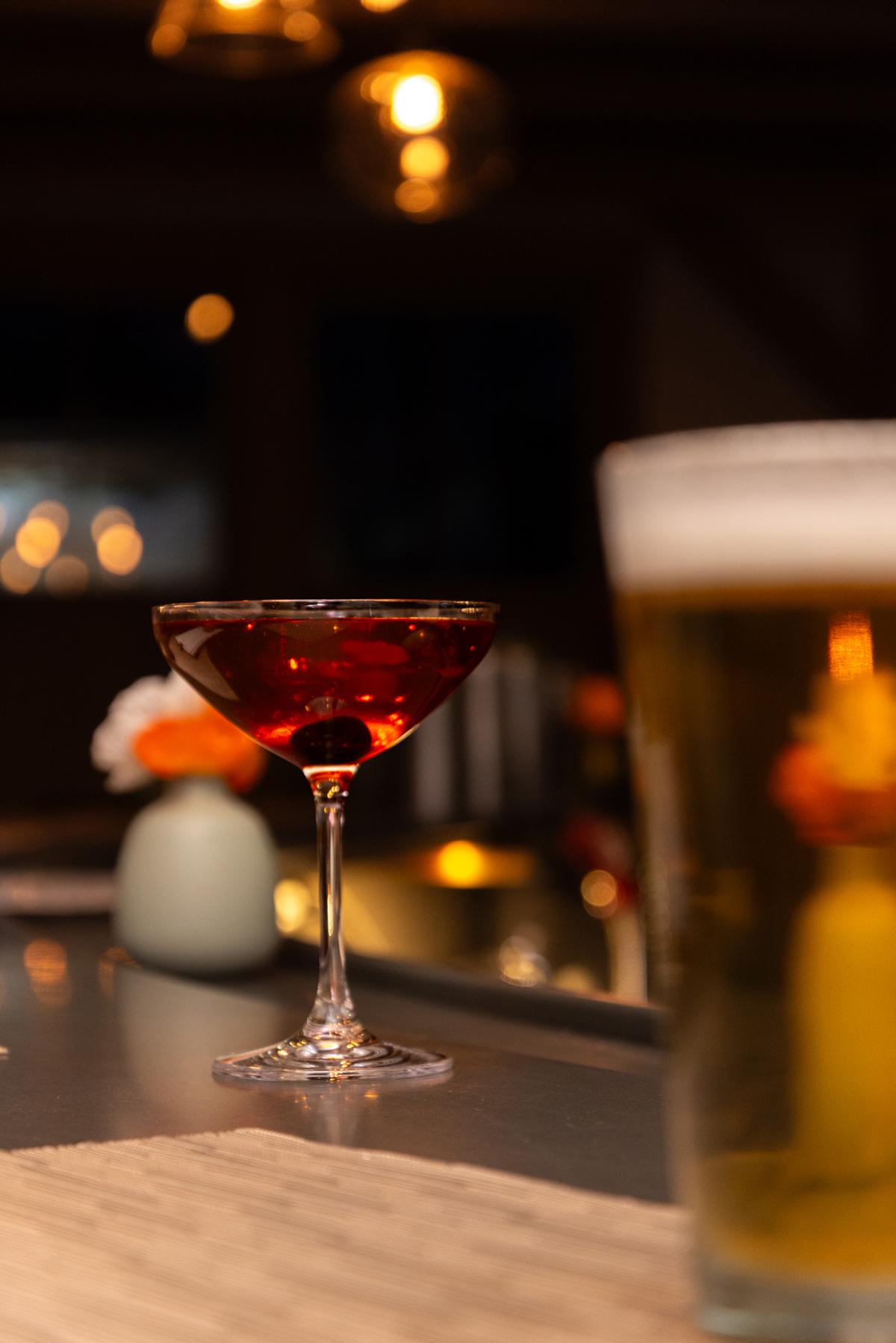 Cocktail with a cherry and a beer glass on a dimly lit bar counter.