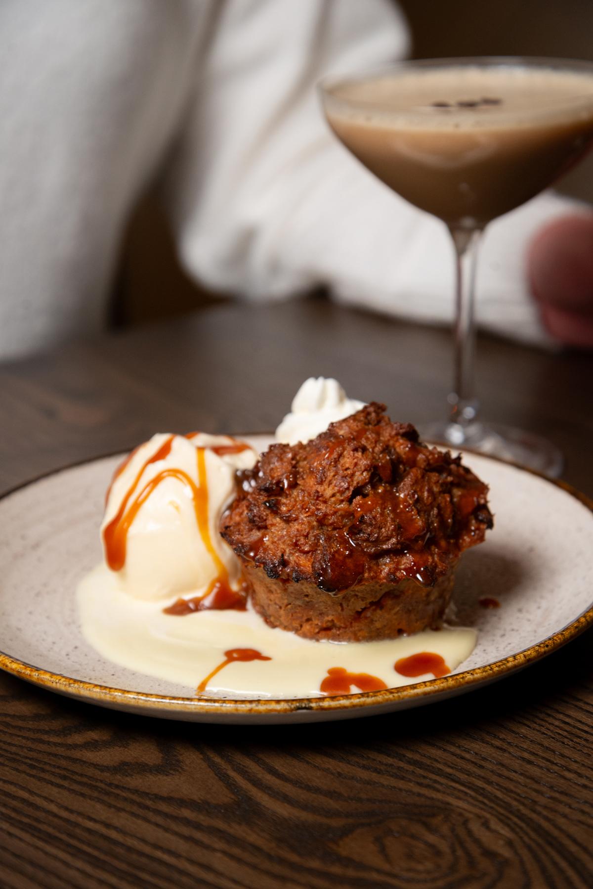 Bread pudding with caramel sauce and ice cream, near a cocktail on wooden table.