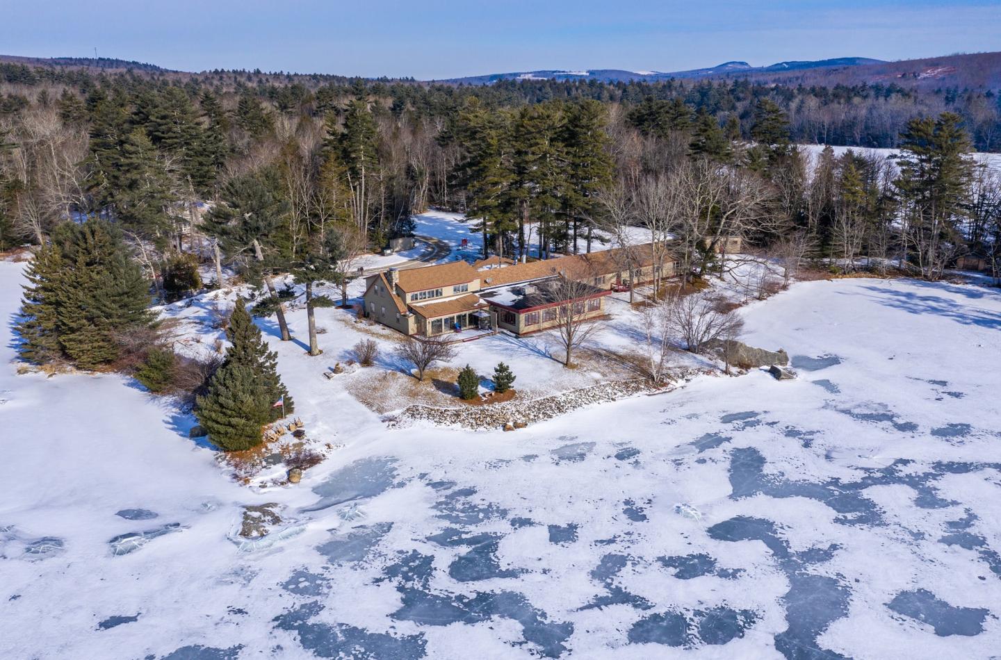 Snowy landscape with house by frozen lake, surrounded by trees.