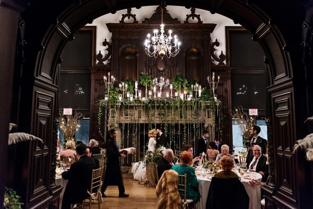 Elegant dining hall with chandeliers, guests seated at tables, and lush greenery decor.