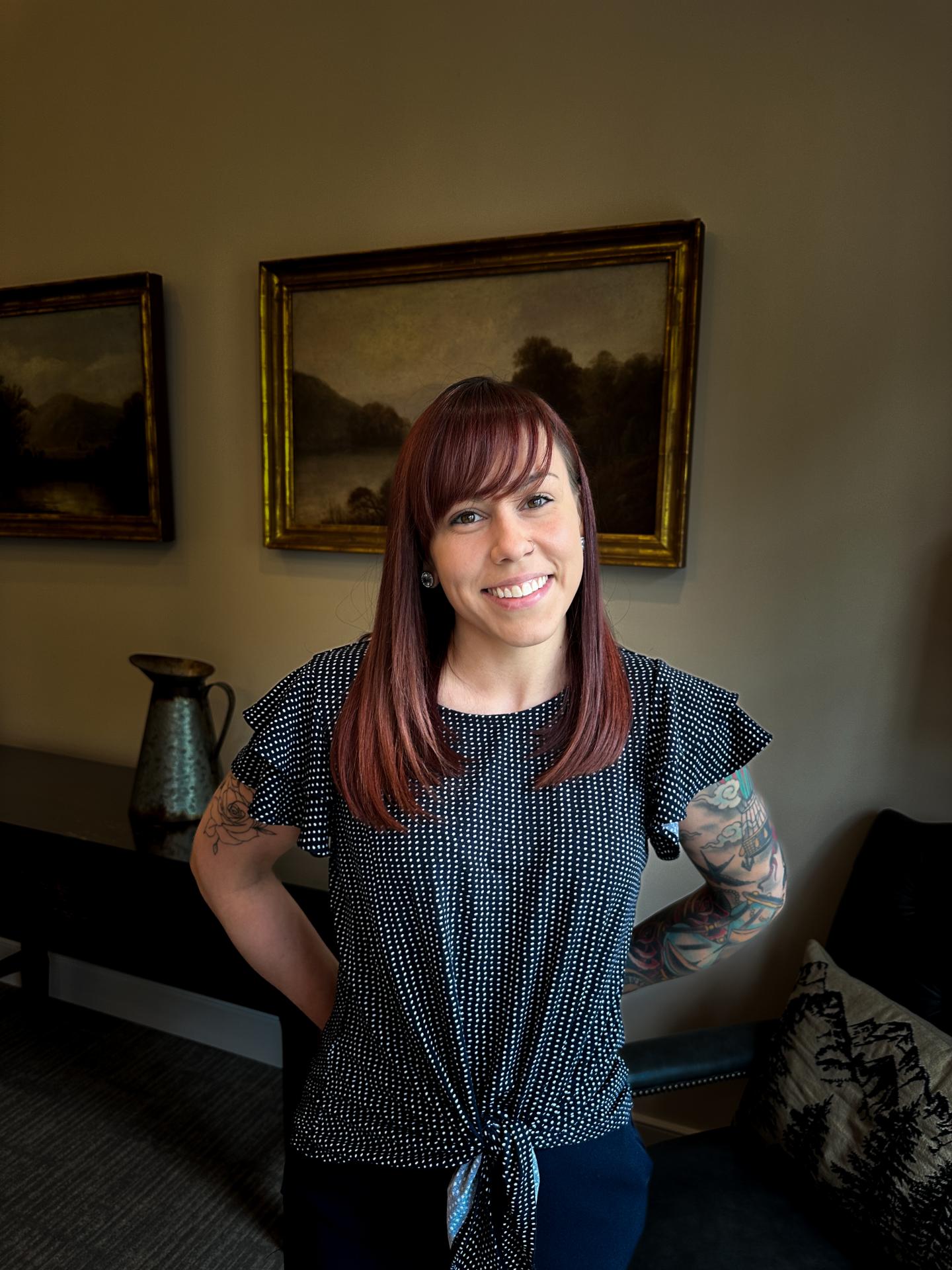 Smiling woman with red hair and tattoos stands in a room with paintings.