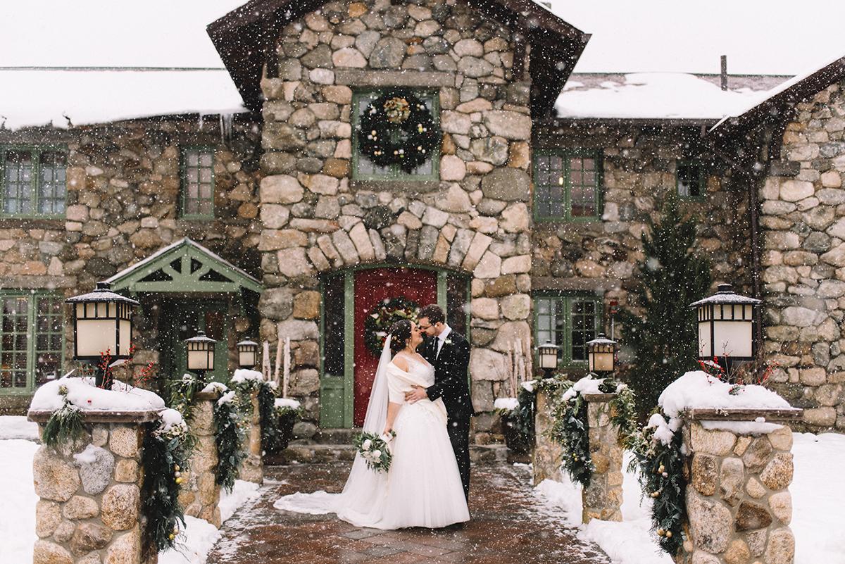 Bride and groom kiss in front of snowy stone building with wreath.