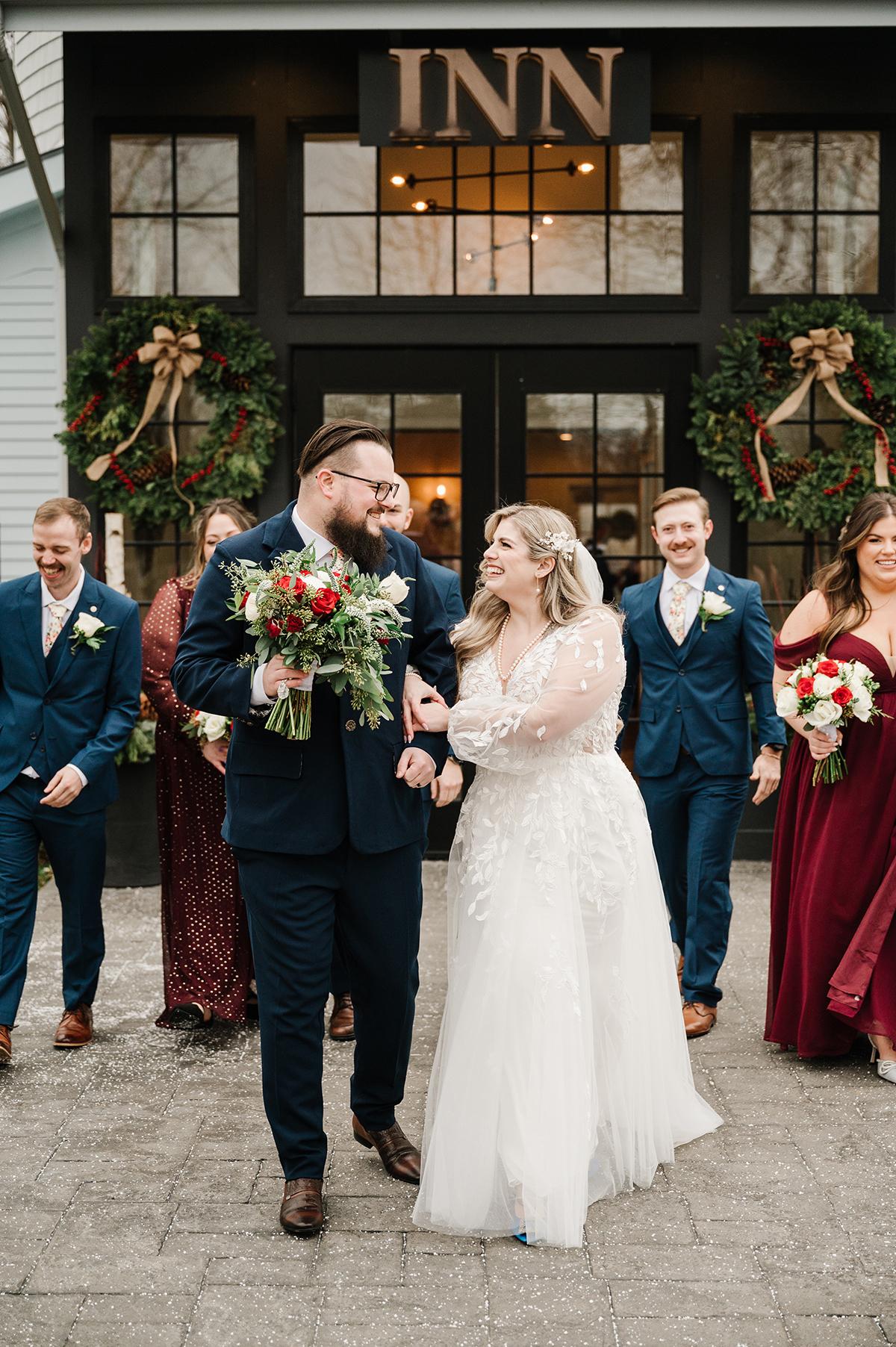 Bride and groom smiling, walking with bridal party in front of an inn.
