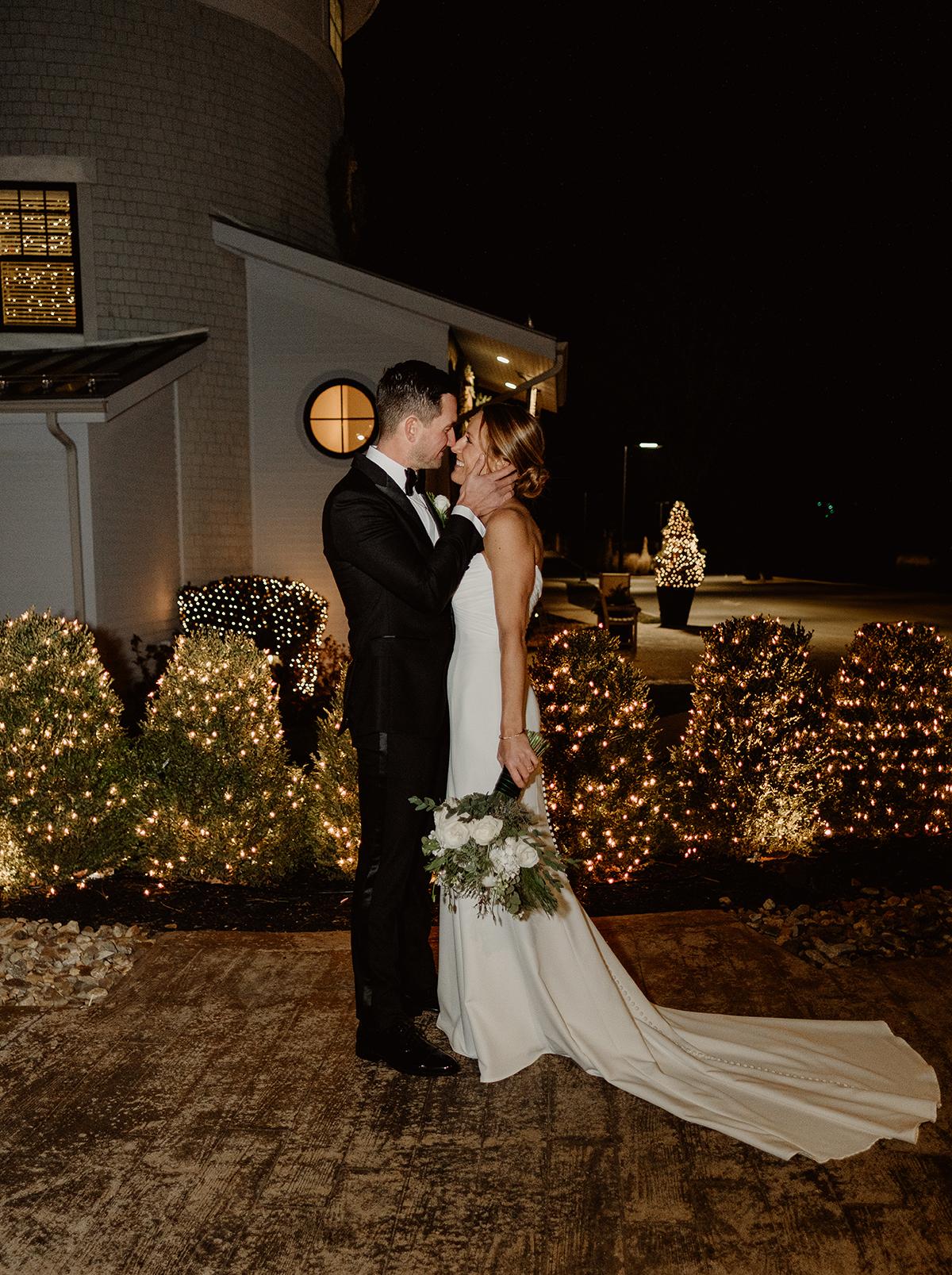 Bride and groom embrace at night, surrounded by twinkling lights.