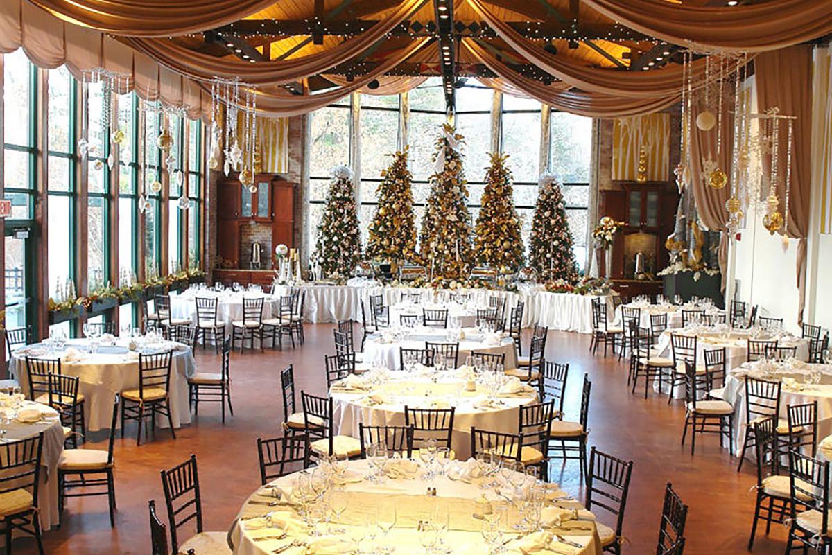 A festive banquet hall with decorated Christmas trees and elegantly set round tables.