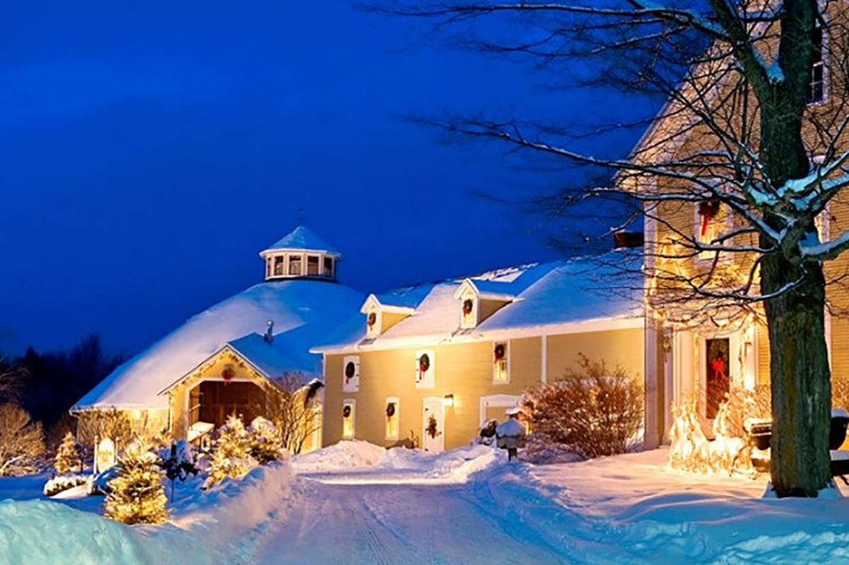 Snow-covered house and barn lit warmly against a deep blue evening sky.