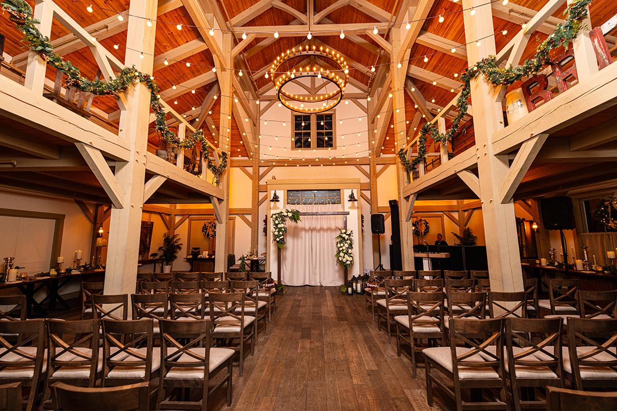 Rustic wedding venue with wooden beams and aisle, decorated with greenery and lights.