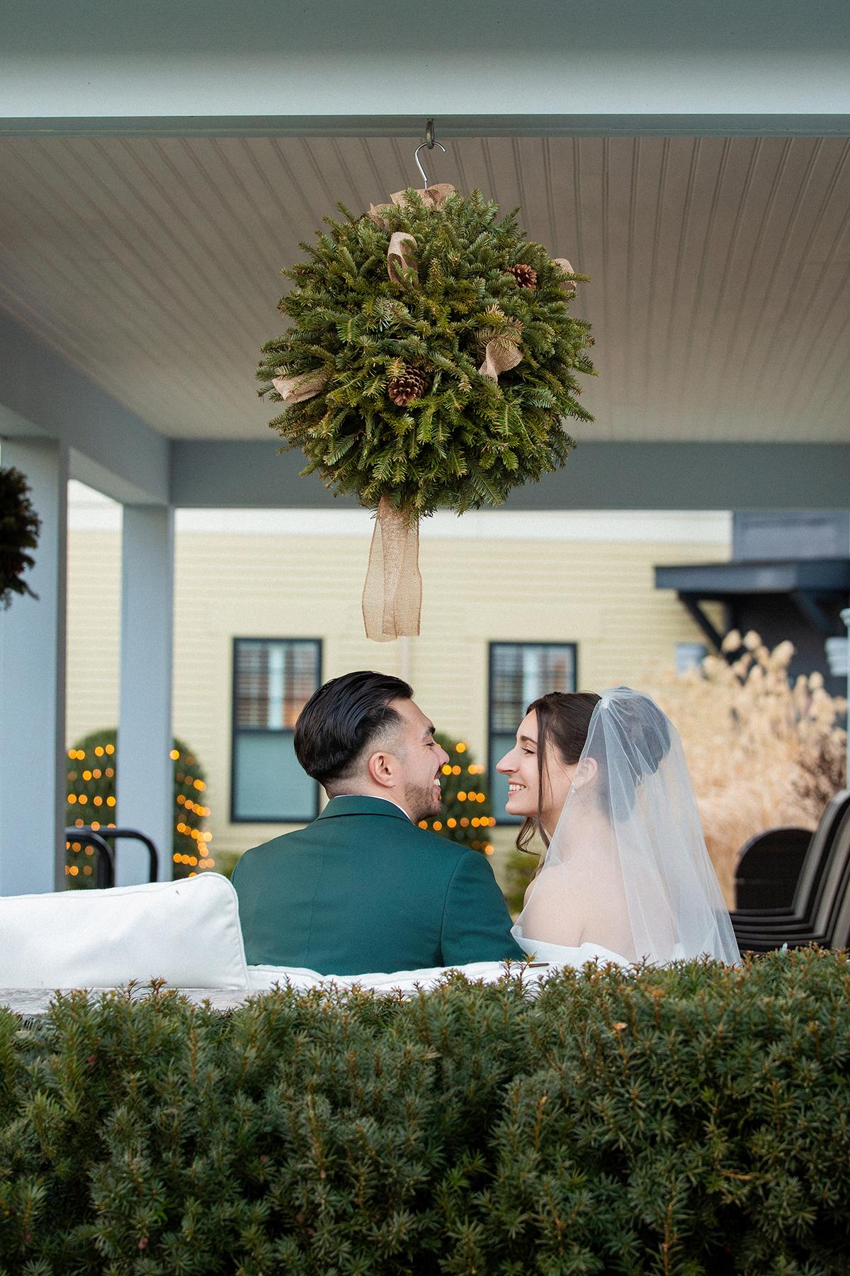 Couple in wedding attire sitting under a decorated porch, exchanging a joyful glance.