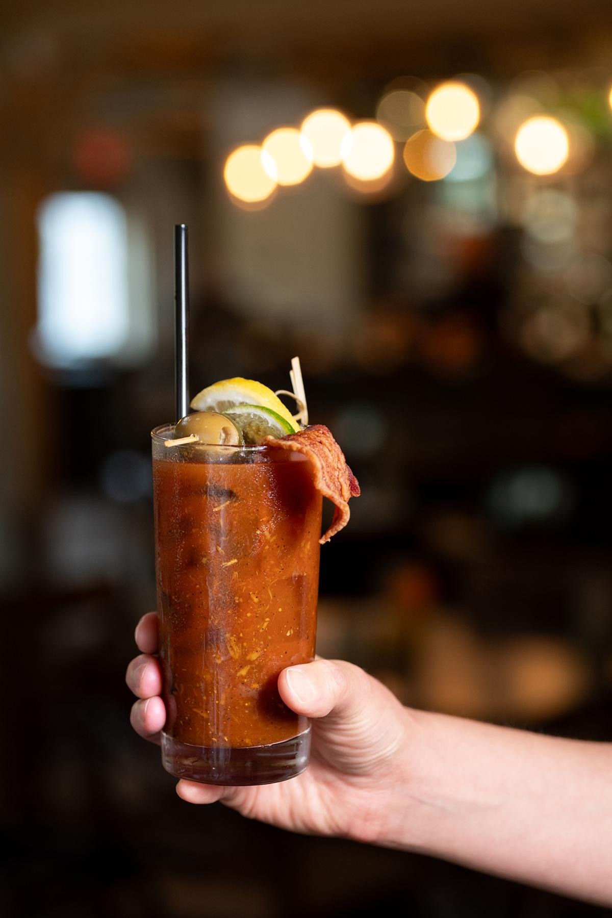 Bloody Mary in a glass garnished with lemon, pickles, and bacon, held by a hand.