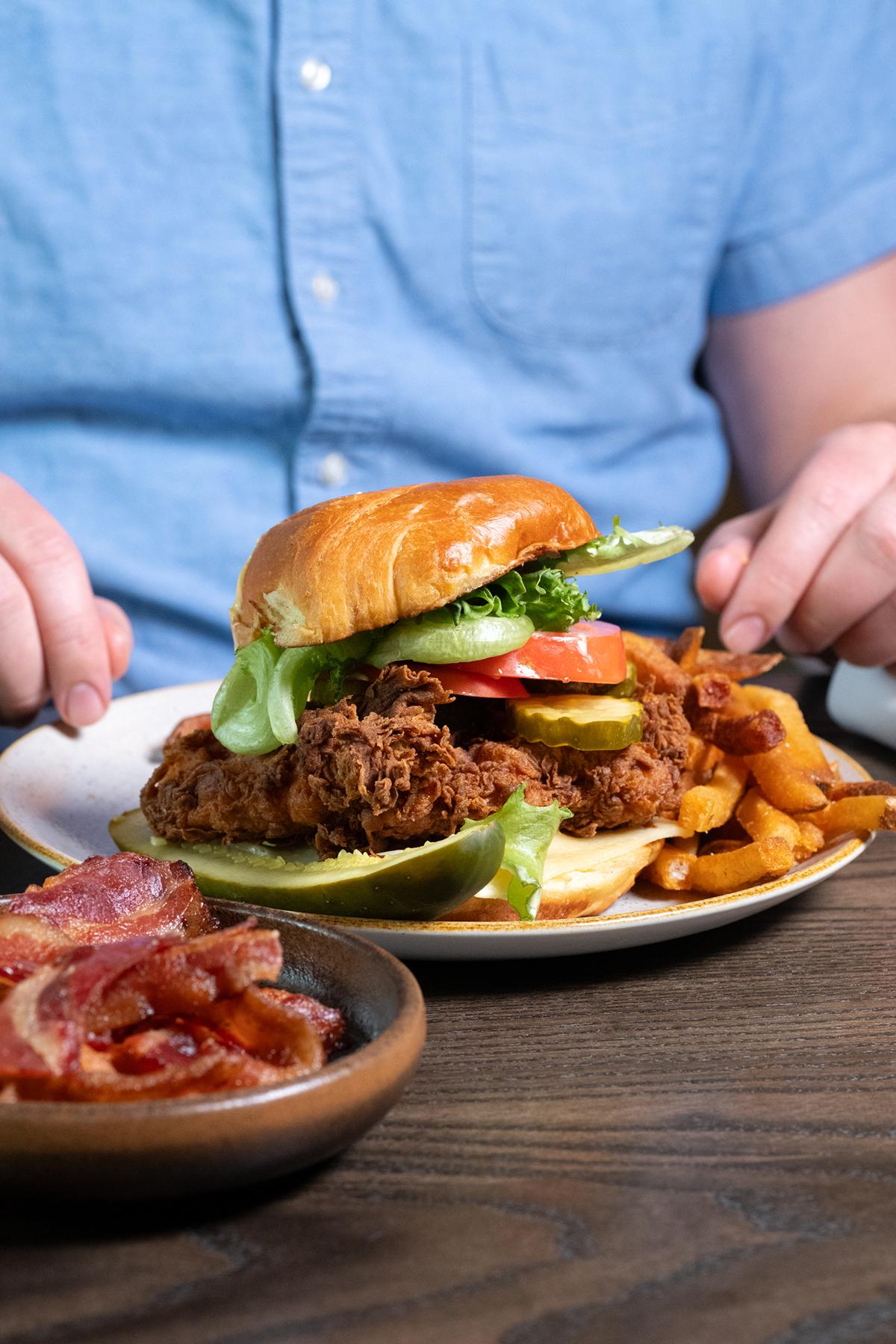 Crispy chicken sandwich with fries on a plate, bacon in a bowl nearby.