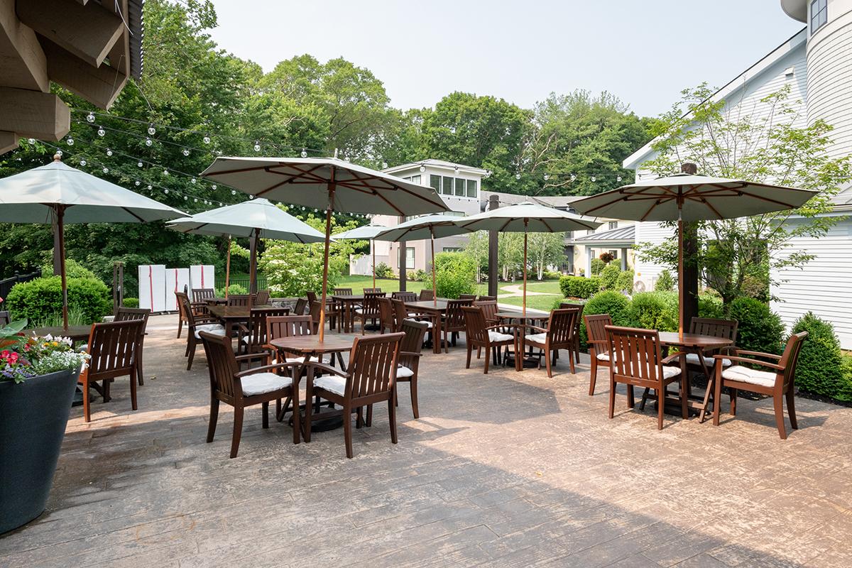 Outdoor patio with wooden tables, chairs, and open umbrellas surrounded by greenery.