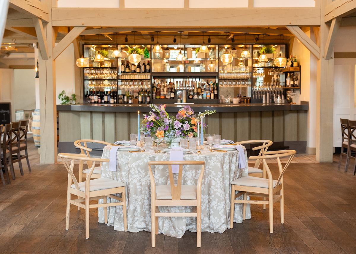 Elegant dining setup with floral centerpiece, wooden chairs, and stocked bar in background.