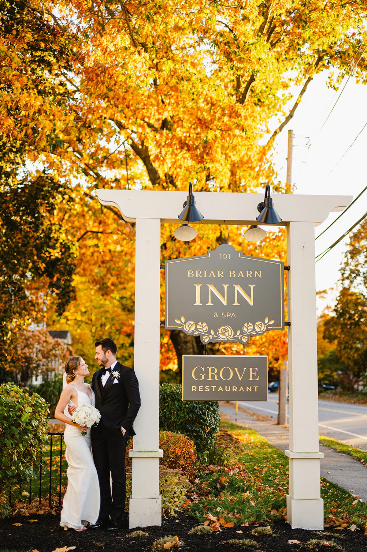 Bride and groom stand under inn sign with autumn leaves.