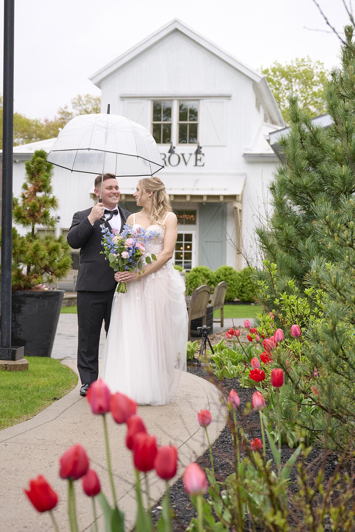 Bride and groom under an umbrella, standing on a garden path with tulips.