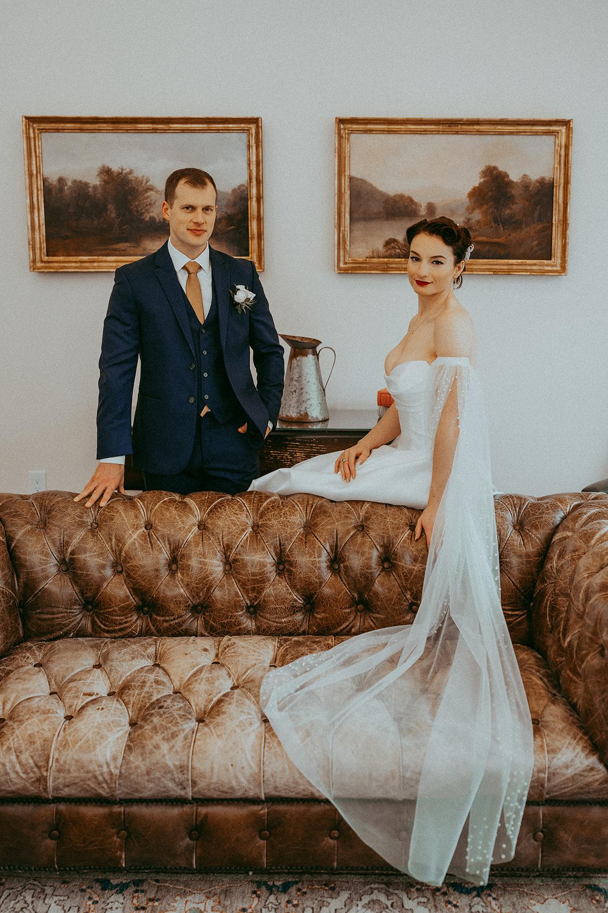 Bride and groom pose beside a vintage couch, with art on the wall behind them.