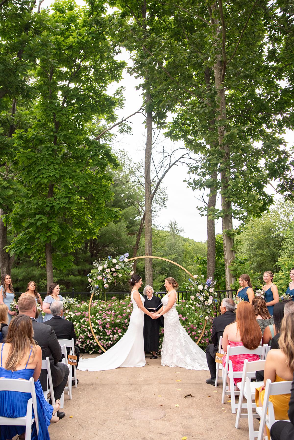 Bride and groom under floral arch in wooded outdoor wedding ceremony.