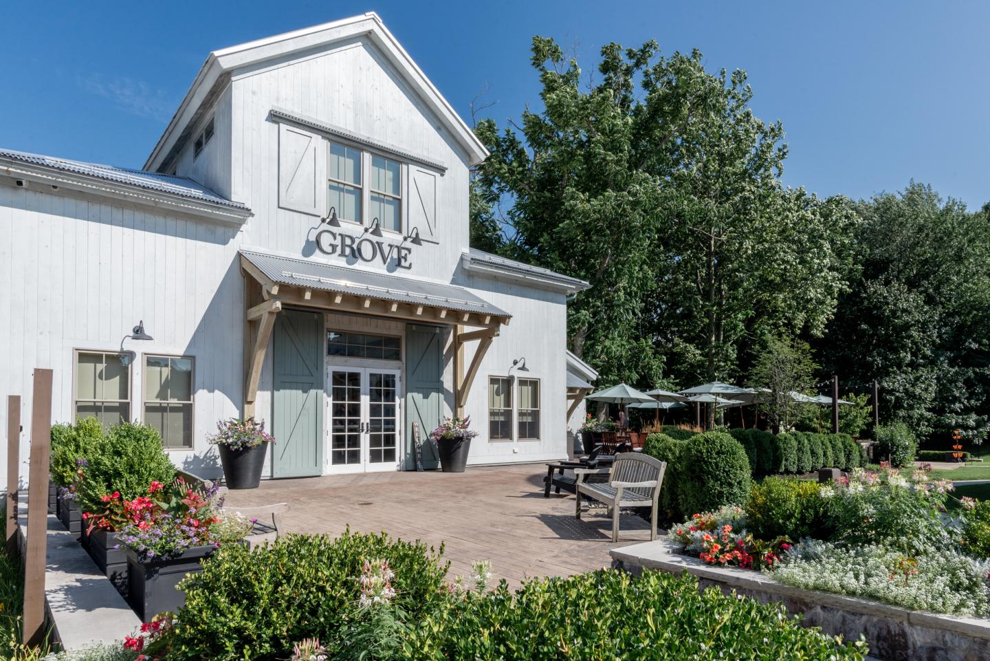 White farmhouse with patio seating, surrounded by lush greenery and colorful flowers.