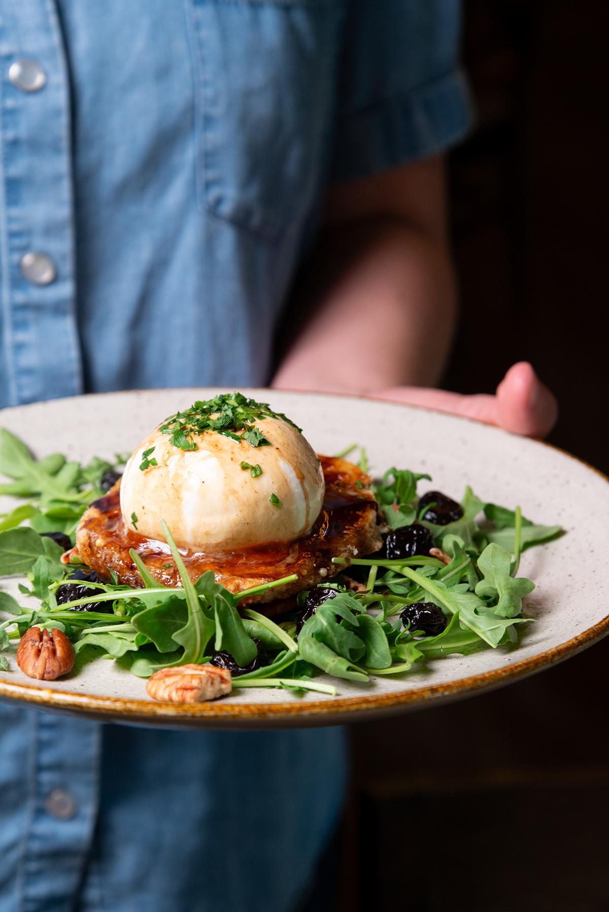 Burrata on arugula salad with nuts and berries, held by a person in a blue shirt.