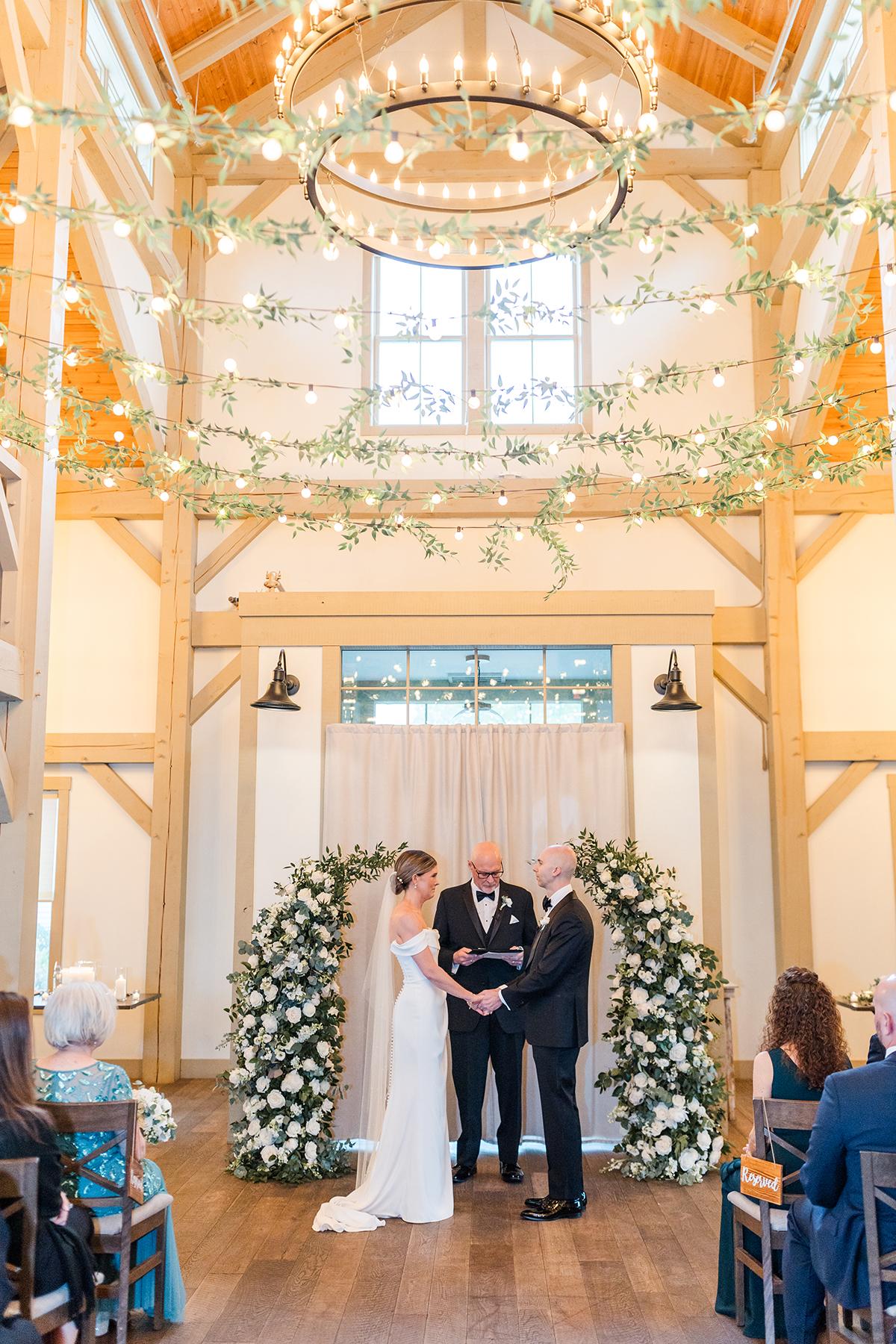Wedding ceremony in a bright hall with floral arch and string lights.