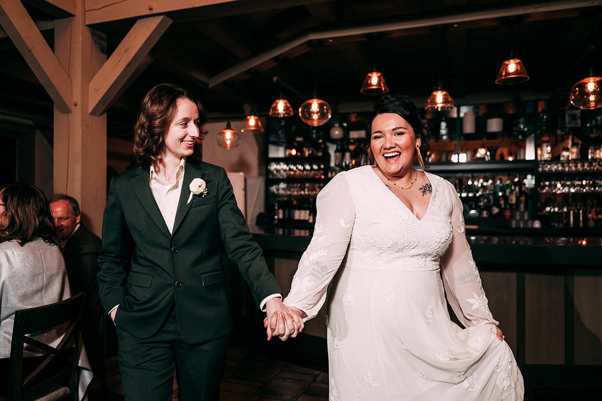 A joyful couple holds hands, dressed in wedding attire with a bar in the background.
