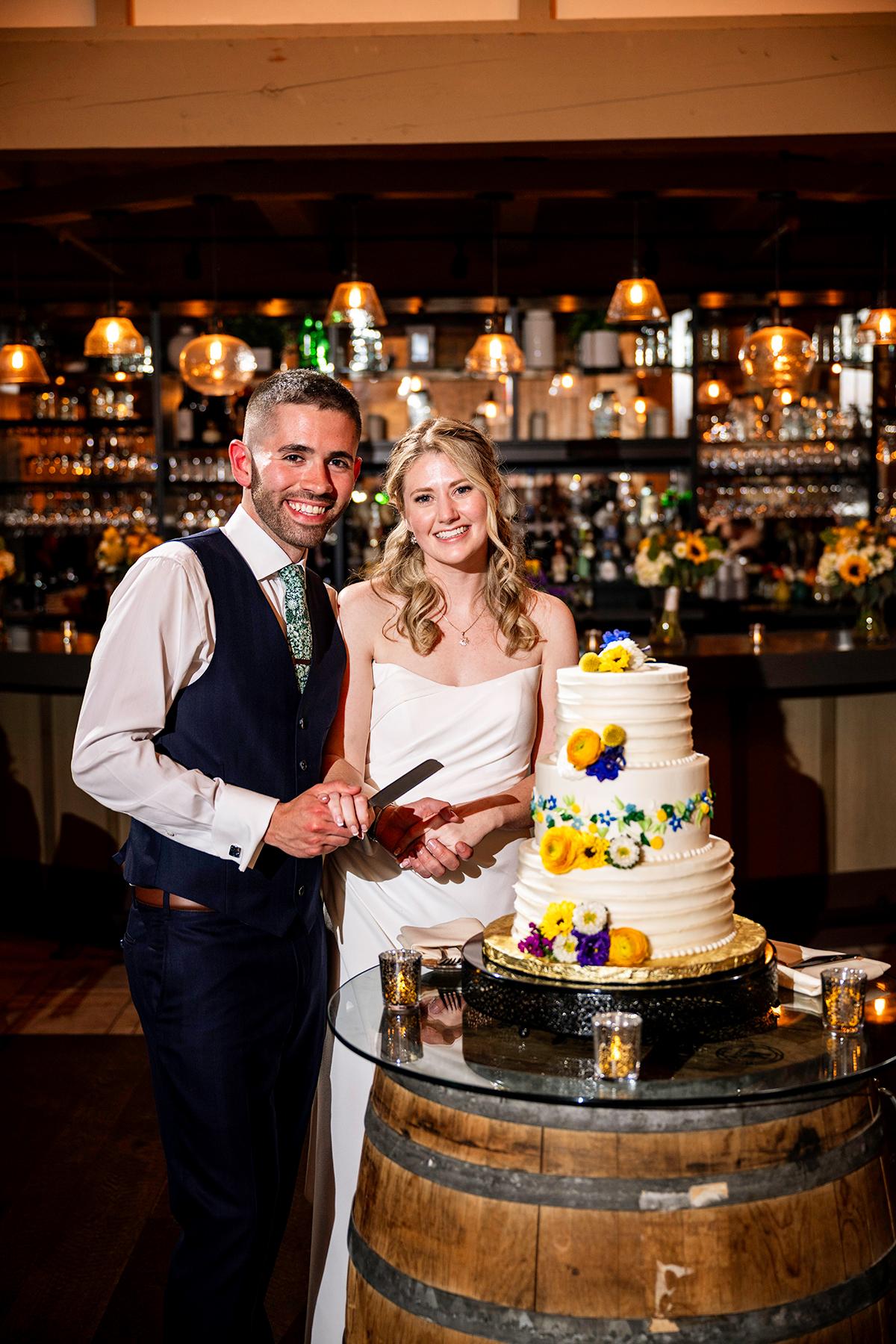 Bride and groom smile, cutting a white floral wedding cake.