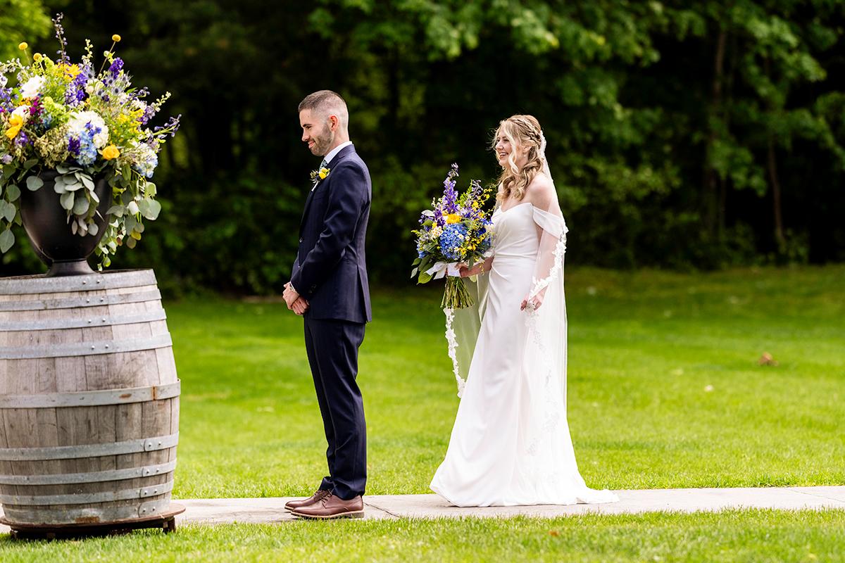 Bride approaches groom outdoors, holding a bouquet, near floral barrel.