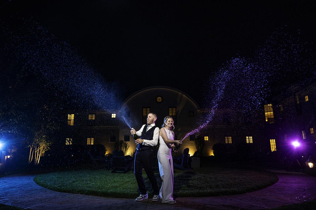 A couple sprays champagne at night, illuminated by lights.
