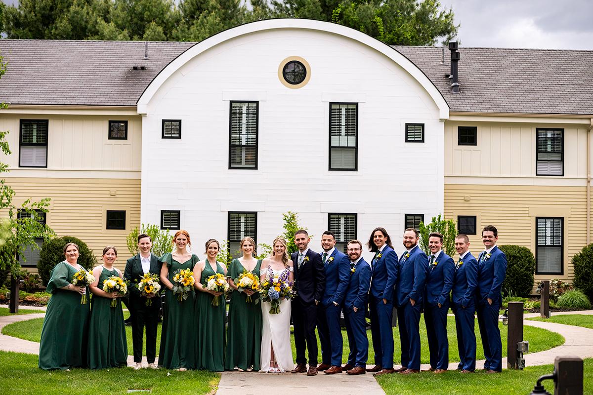 Wedding party in formal attire, posed outside a white building with trees.