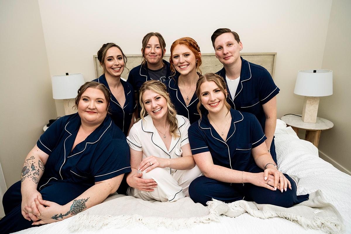 Seven people smiling on a bed, one in white, the rest in navy outfits.