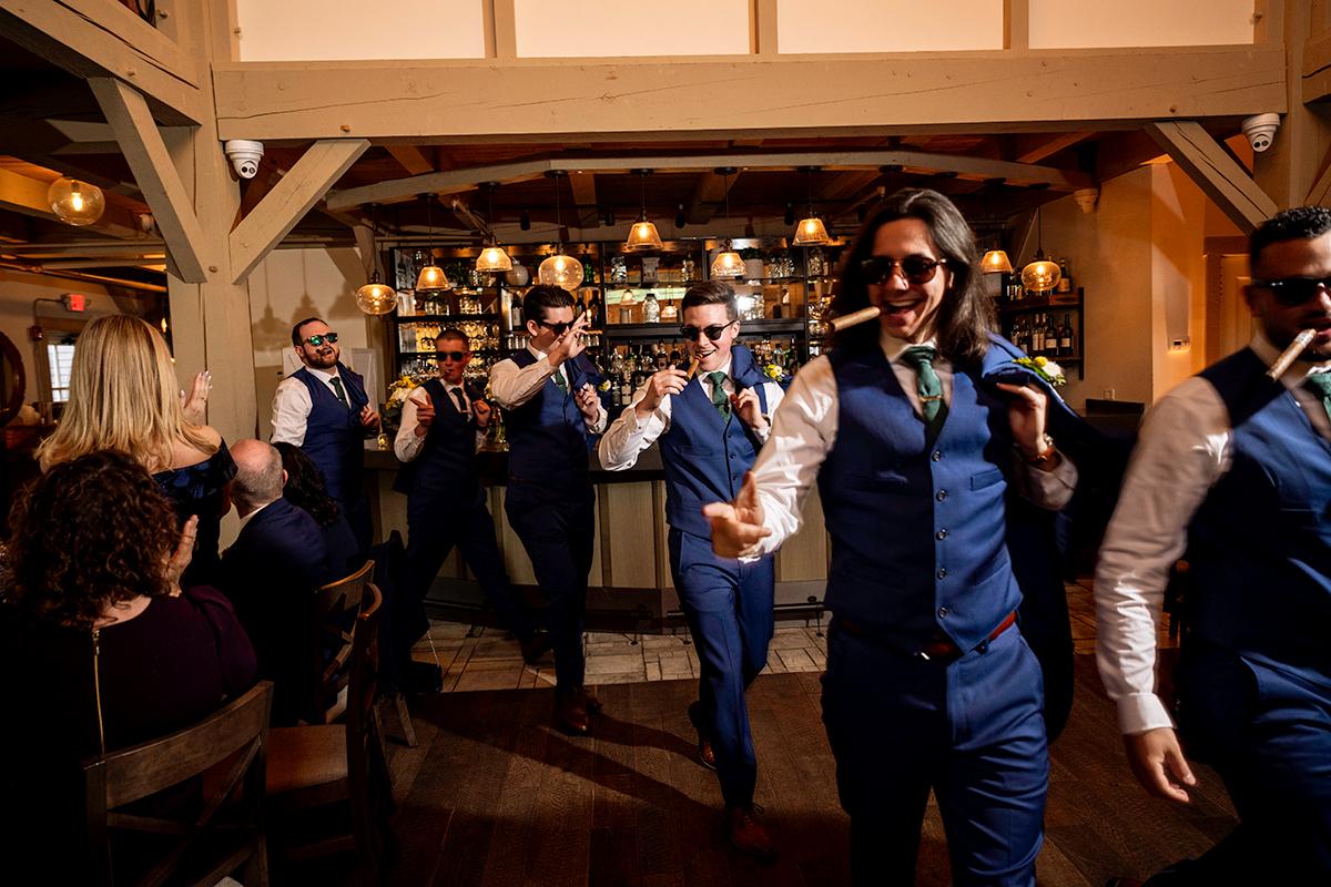 Groomsmen in blue suits dance happily in a rustic venue.