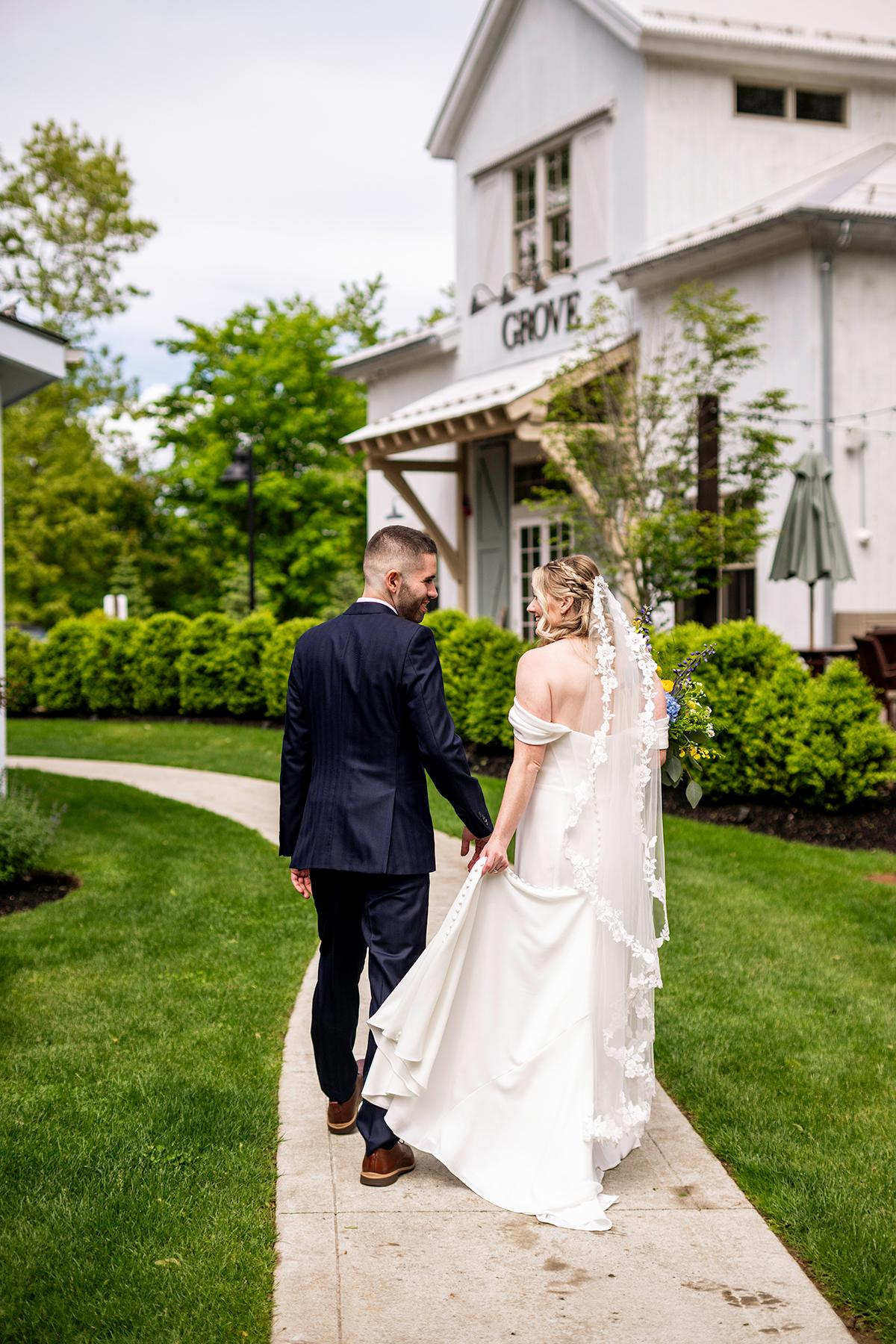 Bride and groom walking on a path, holding hands, near a white building.