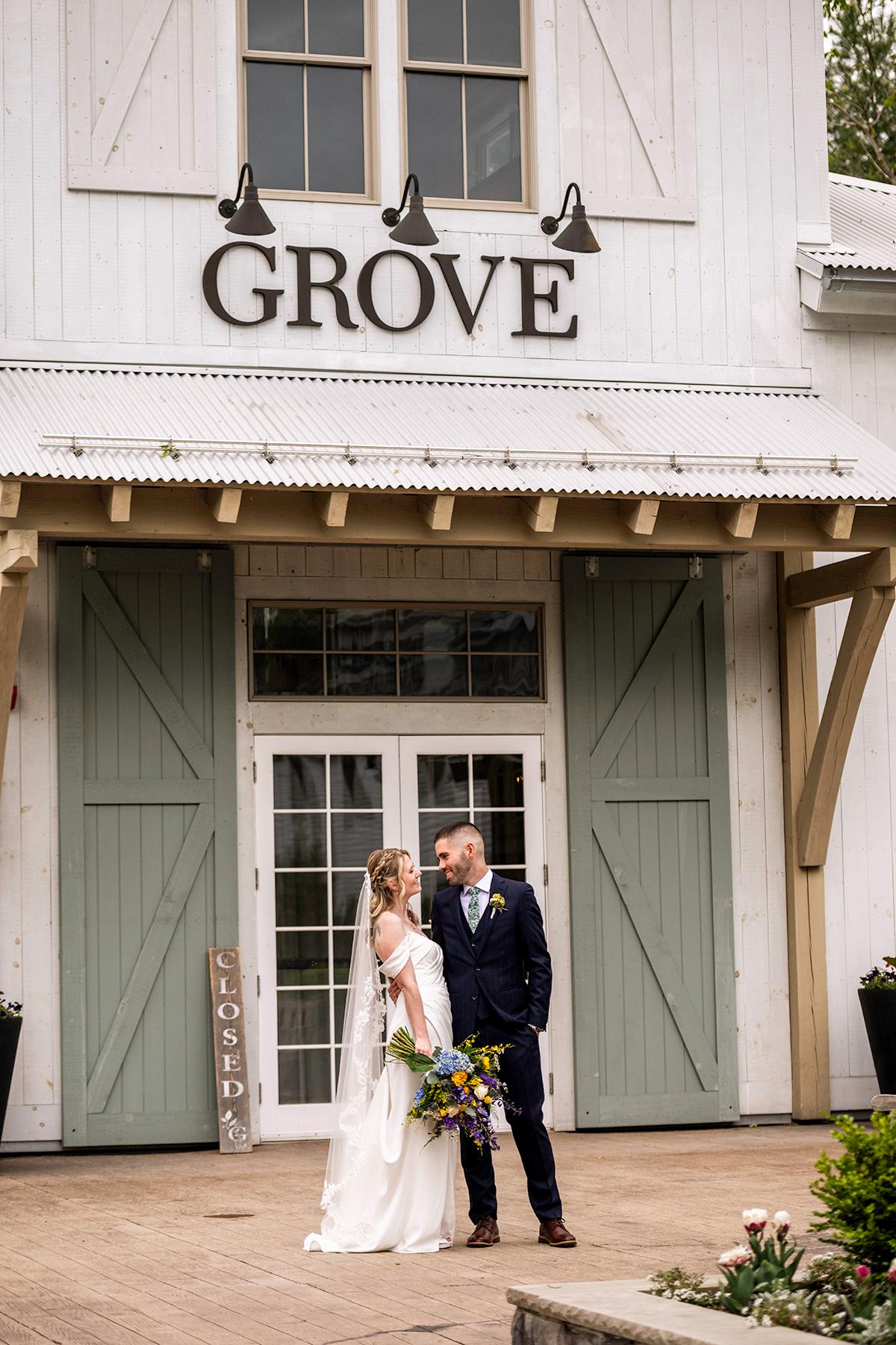 Bride and groom smiling in front of a rustic building labeled "GROVE."