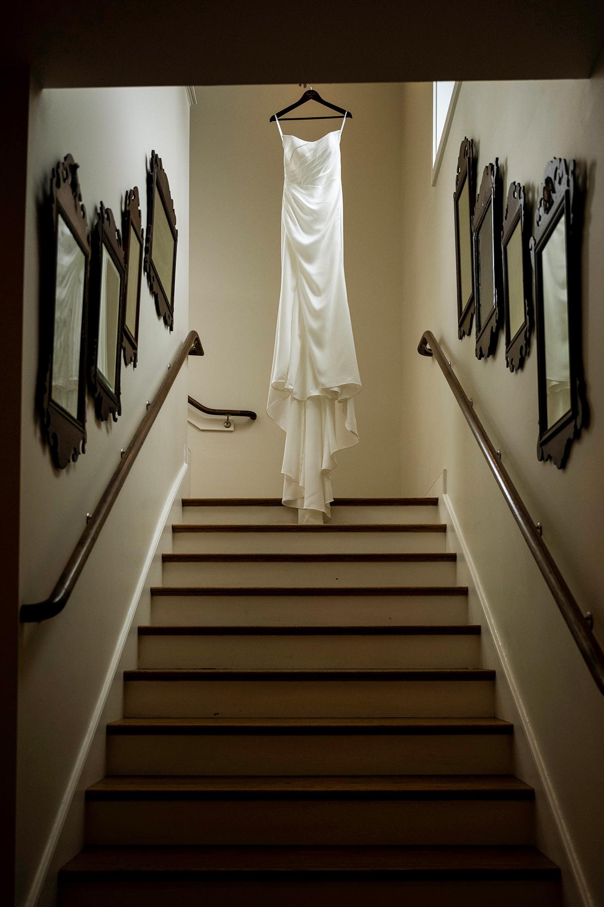 White wedding dress hangs in a stairwell with framed mirrors on the walls.