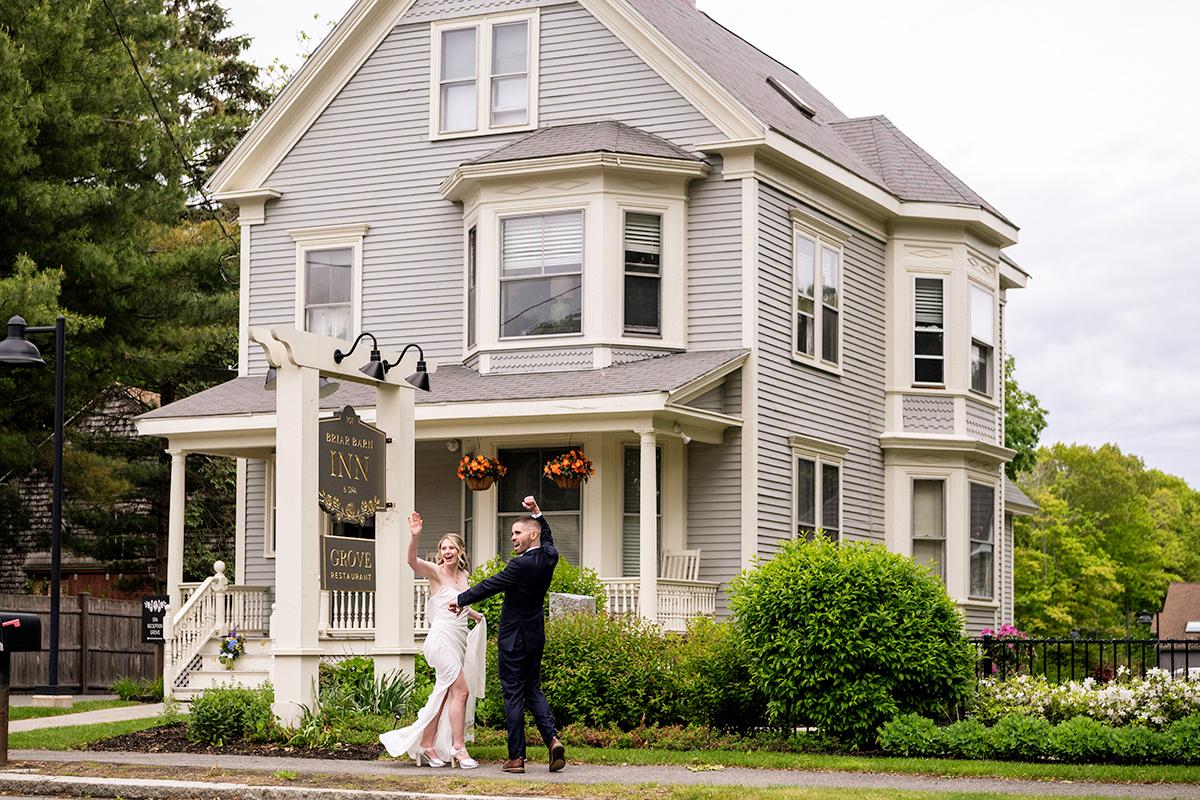 Bride and groom smiling in front of a large, gray Victorian-style house.