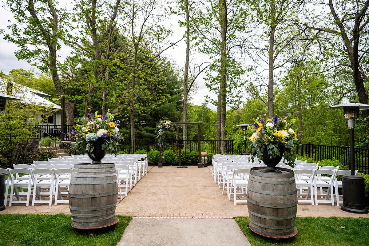 Outdoor wedding setup with rows of white chairs and floral arrangements on wooden barrels.