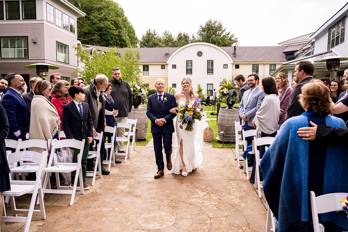 Bride walks down outdoor aisle with older gentleman, guests seated on either side.