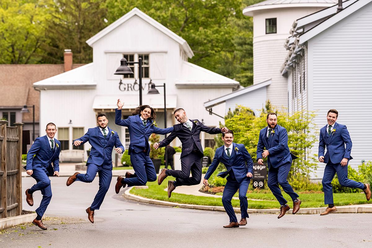 Groomsmen in blue suits jumping with joy outside a white building.