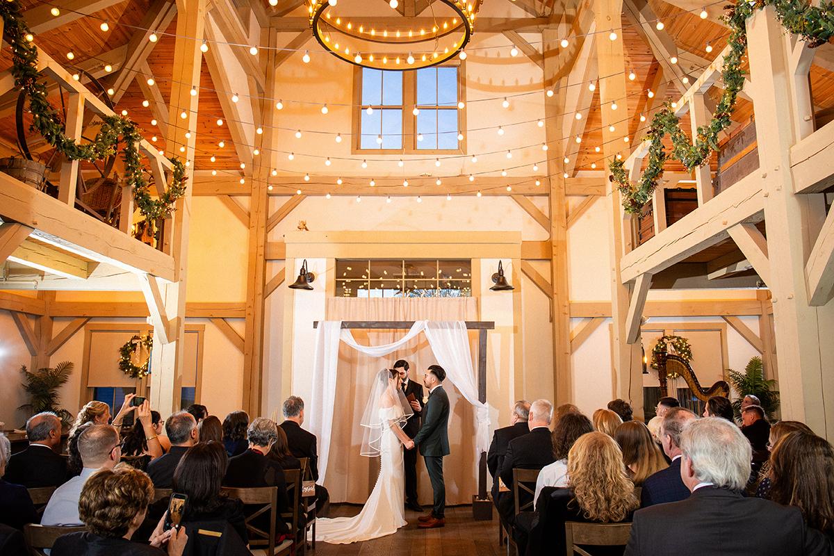 Wedding ceremony in a rustic wooden barn with string lights and guests seated.