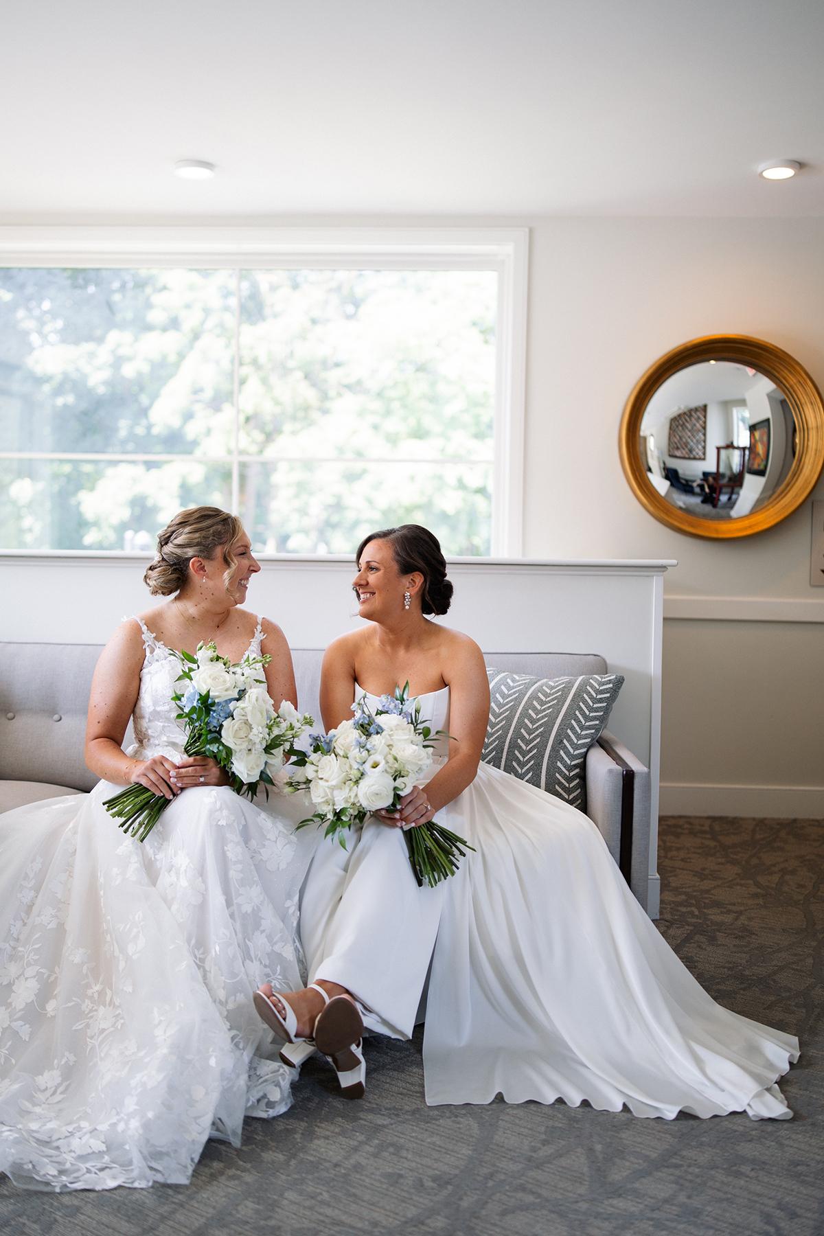 Two brides in white dresses smiling, holding bouquets, seated on a sofa.