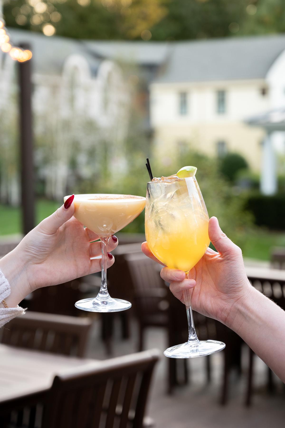Two hands toasting with cocktails on an outdoor patio.