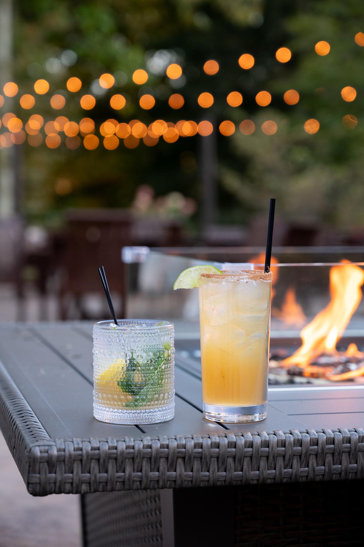 Two cocktails on a table by a fire pit, with string lights in the background.