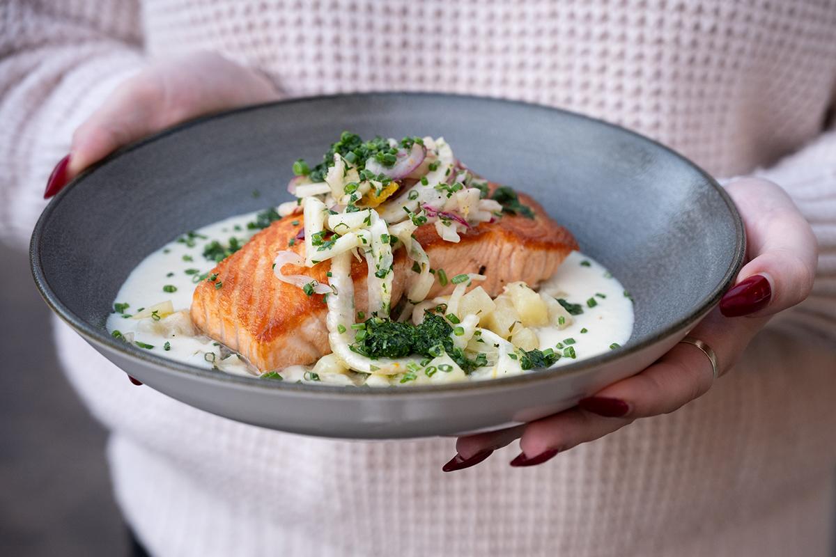 Salmon fillet on creamy sauce, garnished with herbs and noodles, held by a person in a sweater.