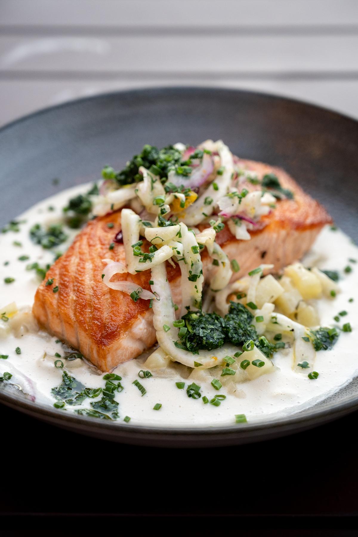 Salmon fillet on white sauce, topped with herbs and vegetables in a dark bowl.