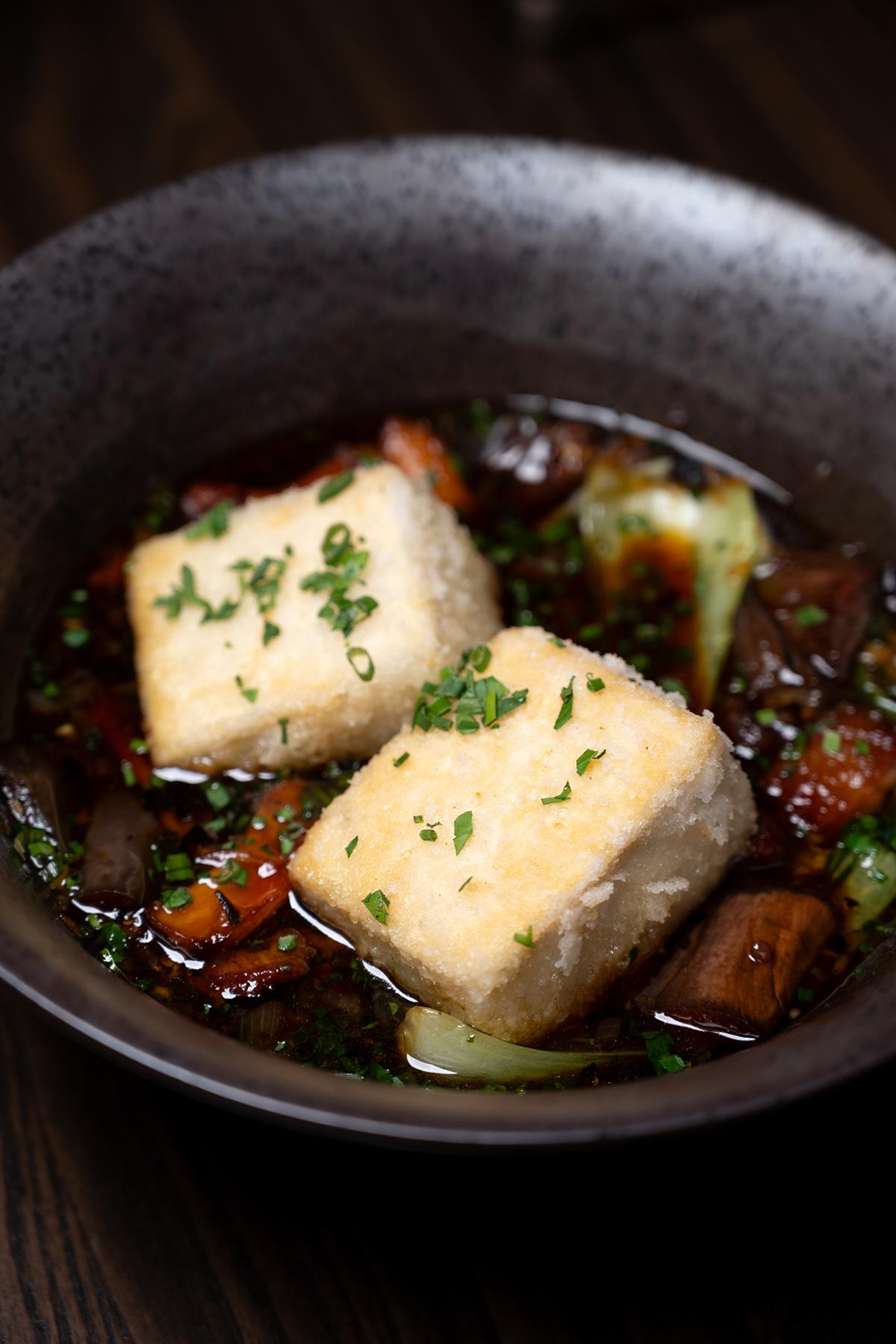 Fried tofu cubes in a rich broth with vegetables, garnished with herbs.