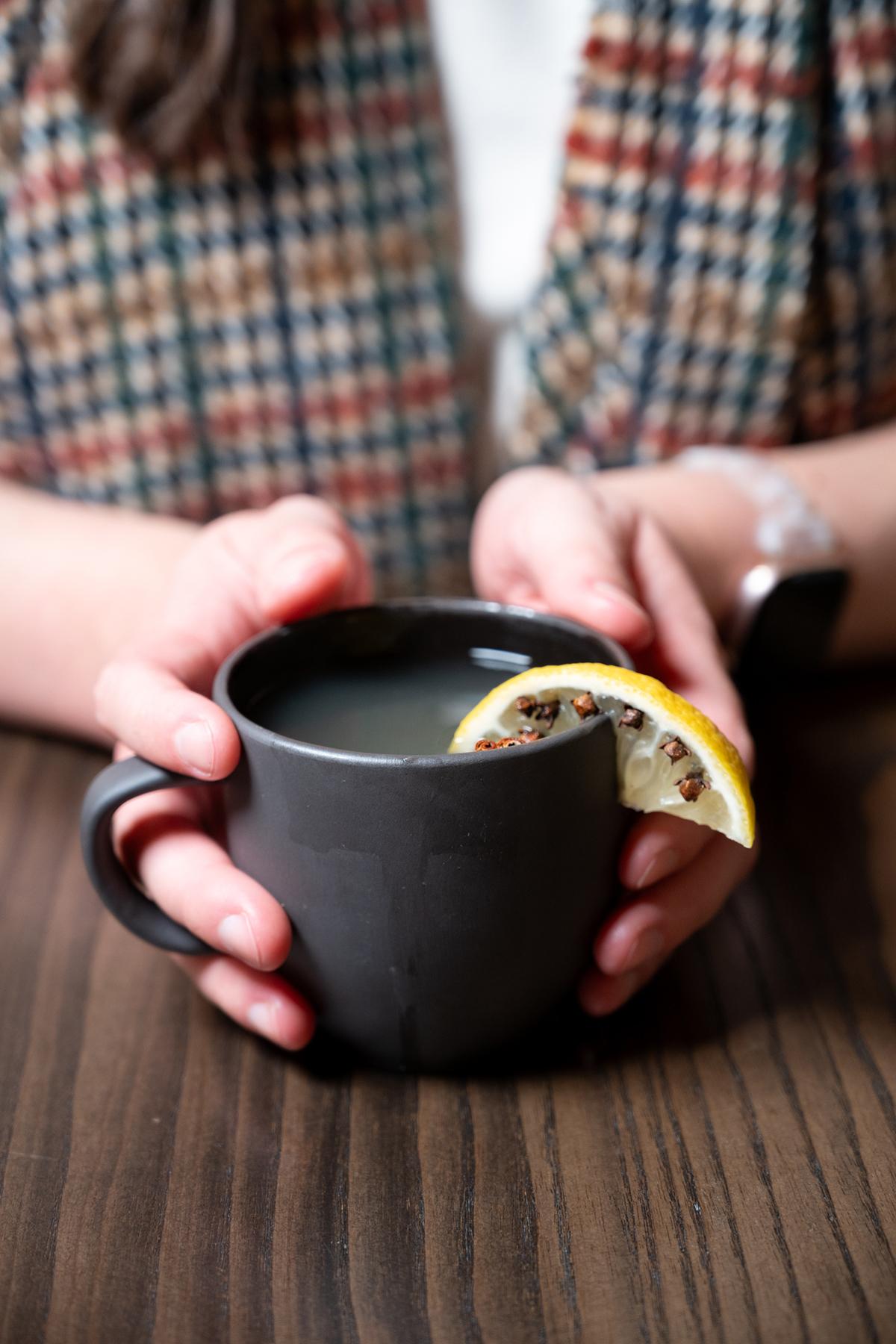 Hands holding a mug with a lemon slice on a wooden table.