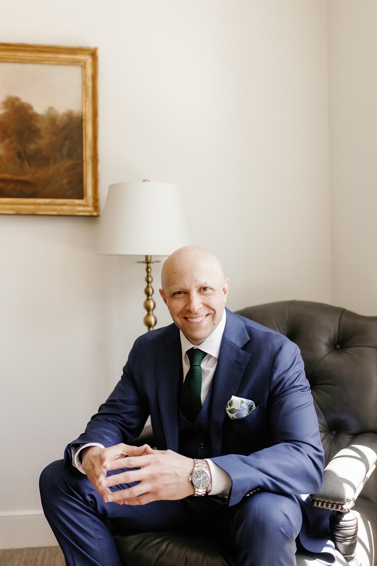 Man in a suit smiling in a cozy room with a painting and lamp.