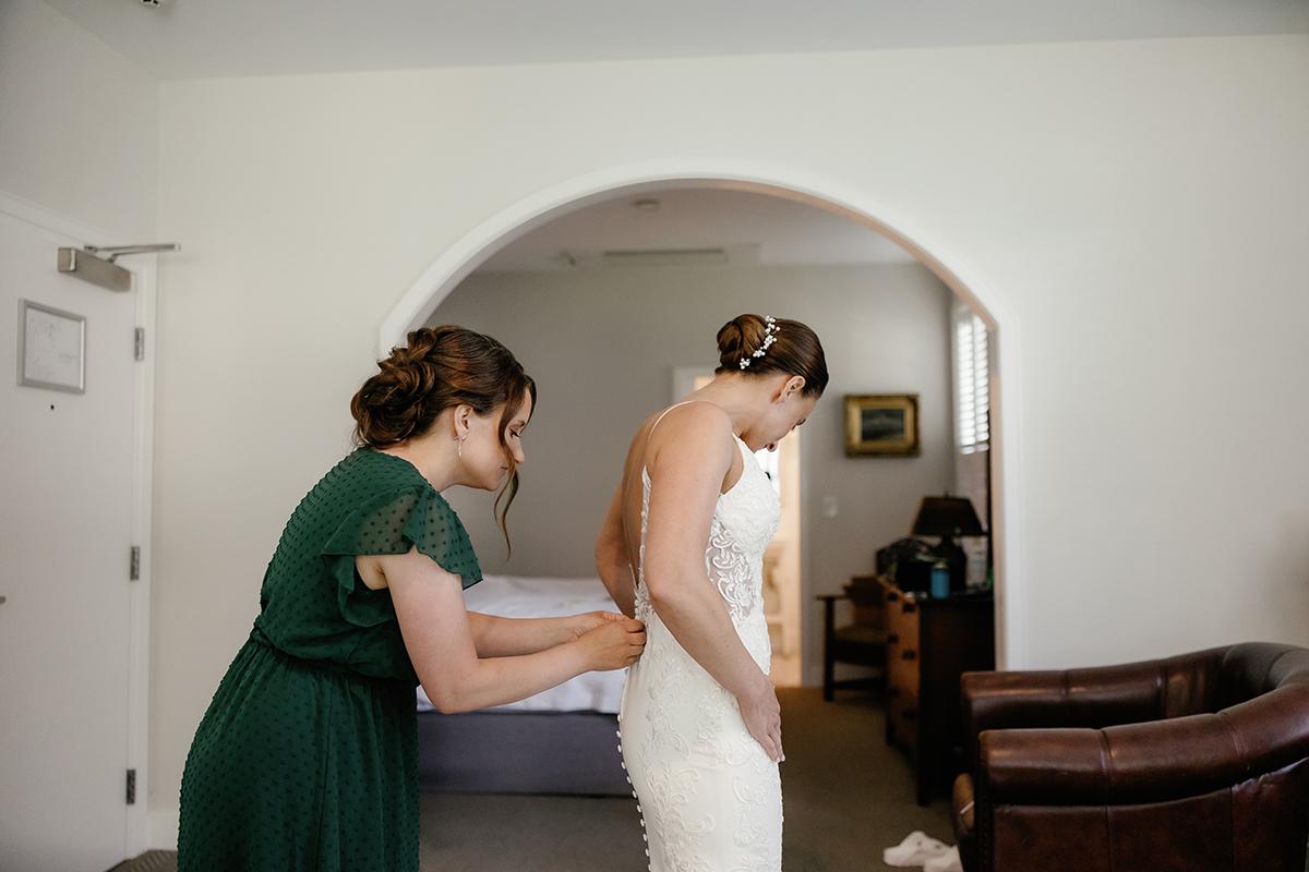 Bride in white dress being helped by woman in green dress in a room.