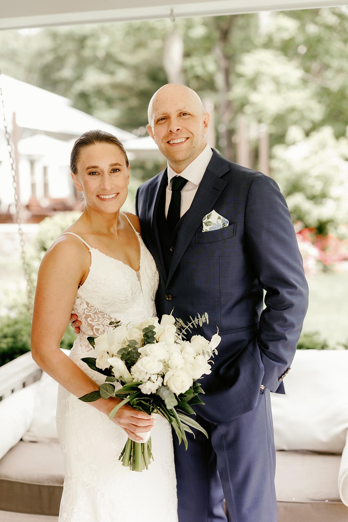 Bride holding a bouquet stands beside the groom in a garden setting.