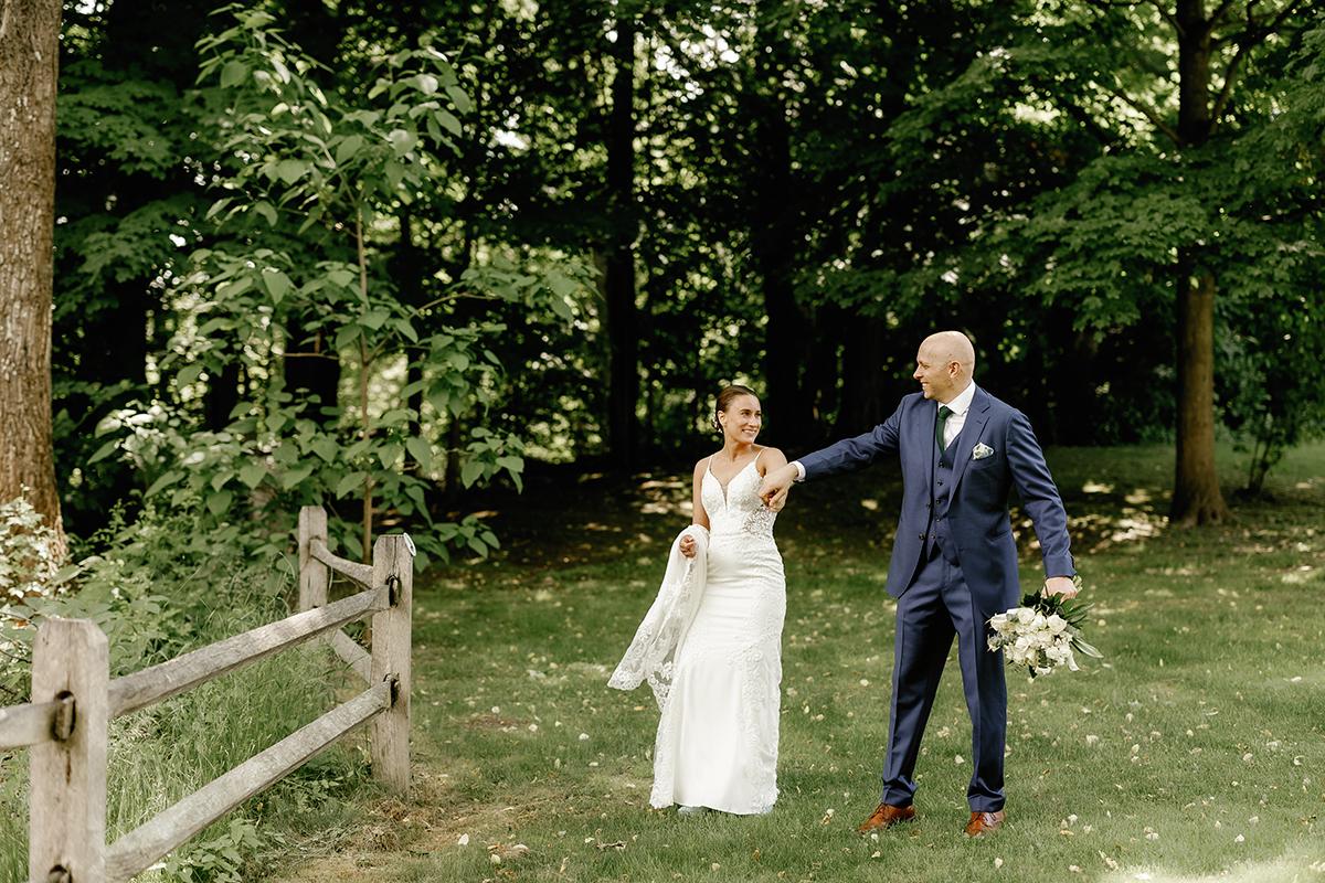 Couple walking in a wooded area, bride in white gown, groom in navy suit holding flowers.