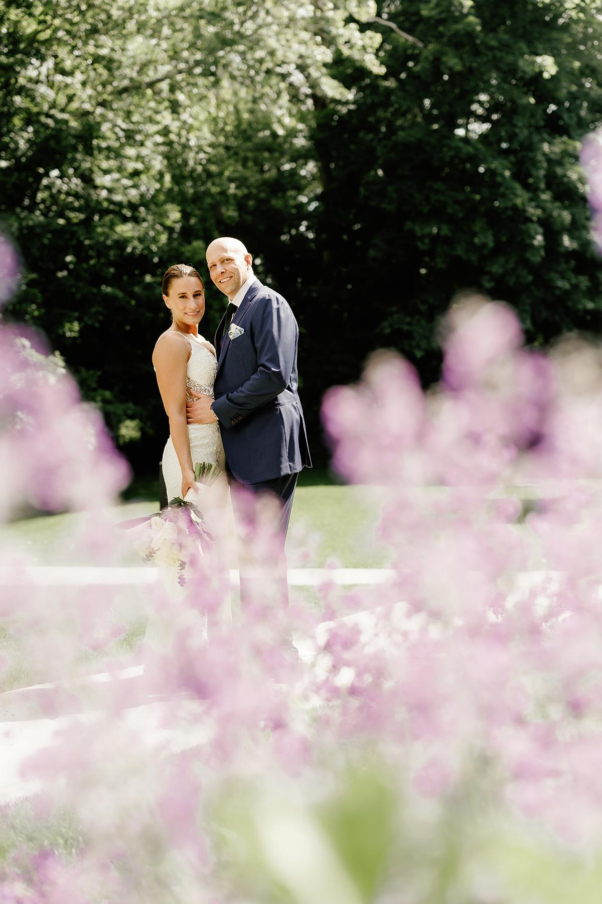 Couple standing in garden, surrounded by purple flowers, with greenery in the background.
